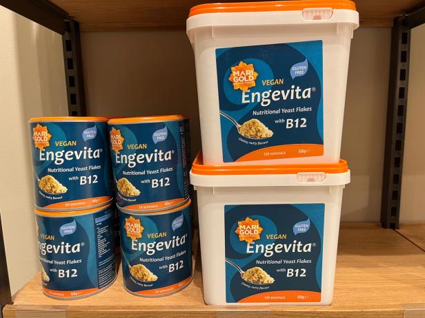 several containers of "Marigold Vegan Engevita Nutritional Yeast Flakes with B12" stacked on a shelf. There are four smaller cans and two larger tubs of the product.