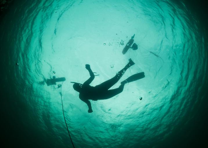 An underwater shot of a diver swimming, with bubbles rising from their equipment. The diver is silhouetted against the turquoise water, with two other divers visible in the background.