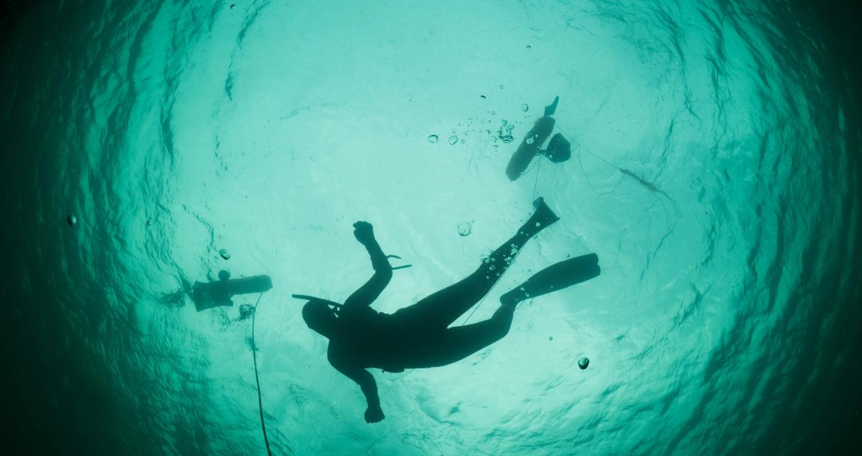An underwater shot of a diver swimming, with bubbles rising from their equipment. The diver is silhouetted against the turquoise water, with two other divers visible in the background.