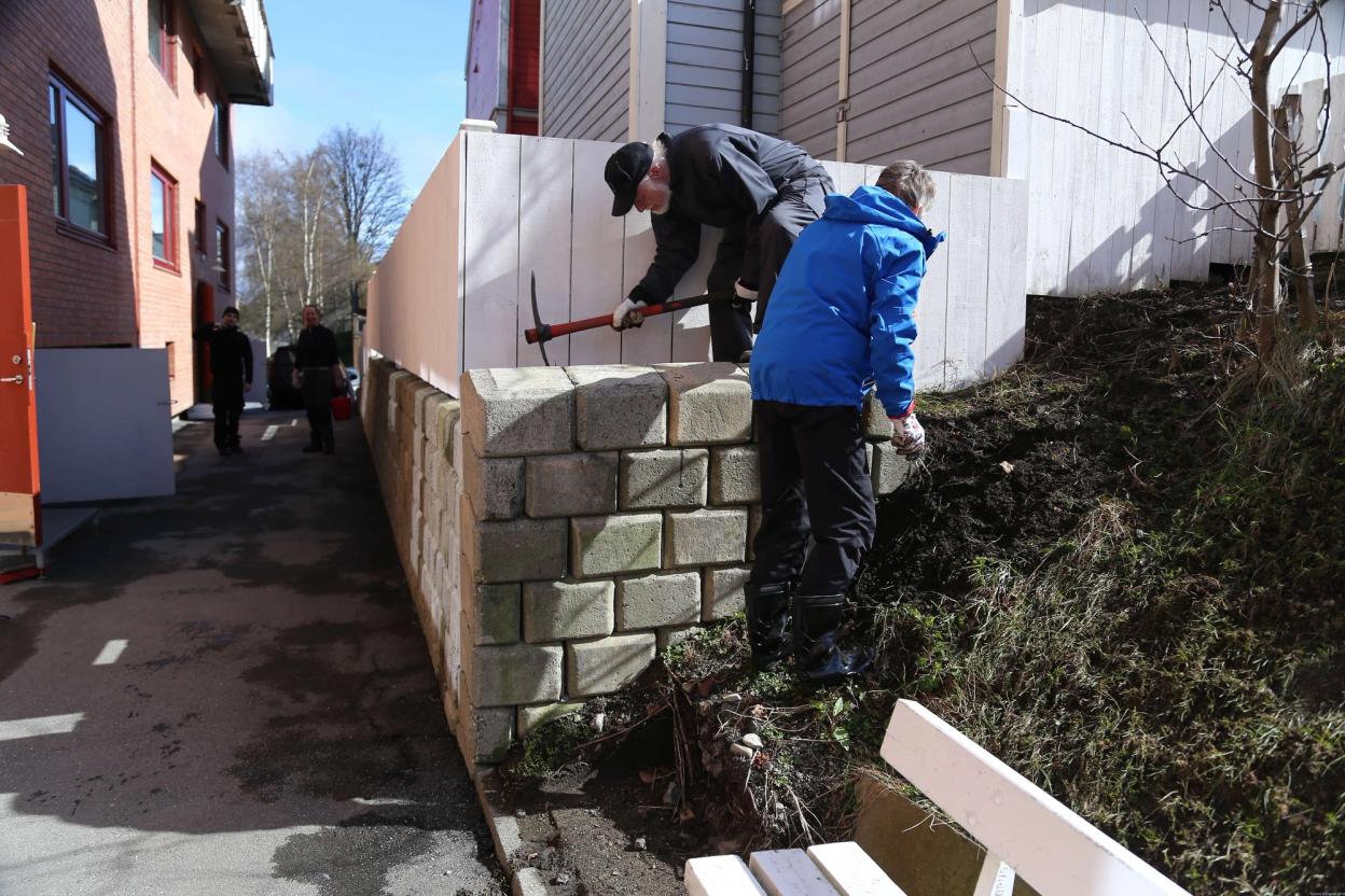 Two volunteers build a retaining wall with large concrete blocks, using a pickaxe to secure the structure.