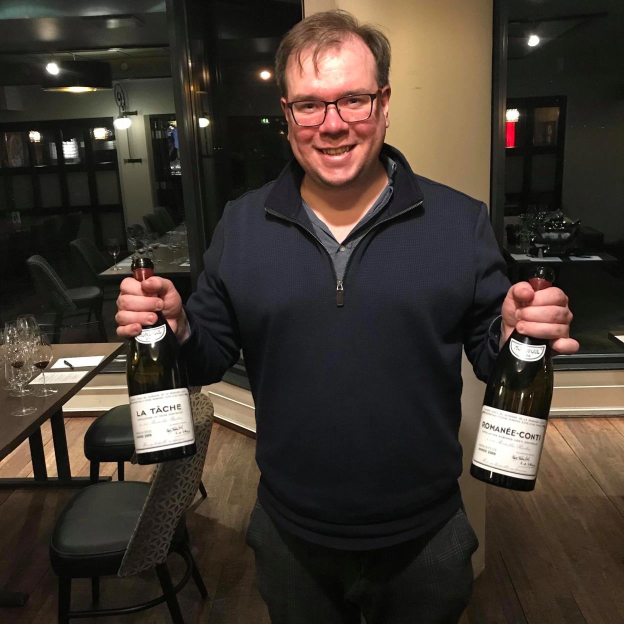 Levi Røstad standing in a dimly lit restaurant, smiling while holding two bottles of fine wine—La Tâche and Romanée-Conti. He is wearing a navy blue sweater over a gray shirt. In the background, tables with wine glasses and chairs are set up, creating an elegant dining atmosphere.