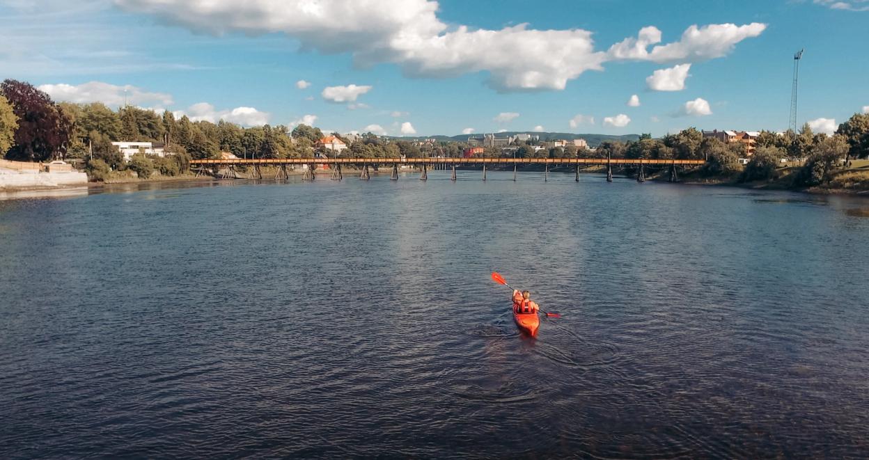 A person kayaking on a calm river under a bright blue sky, with a scenic bridge and lush greenery in the background.