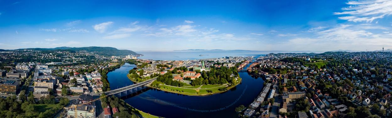 A panoramic view of Trondheim with the Nidelva river winding through the city. The photo captures a vibrant mix of residential buildings, green spaces, and the distant fjord under a clear blue sky with a few wispy clouds.