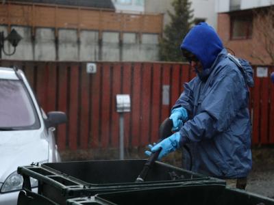 A person in a blue rain jacket and gloves power washes outdoor garbage bins in a residential area on a rainy day.