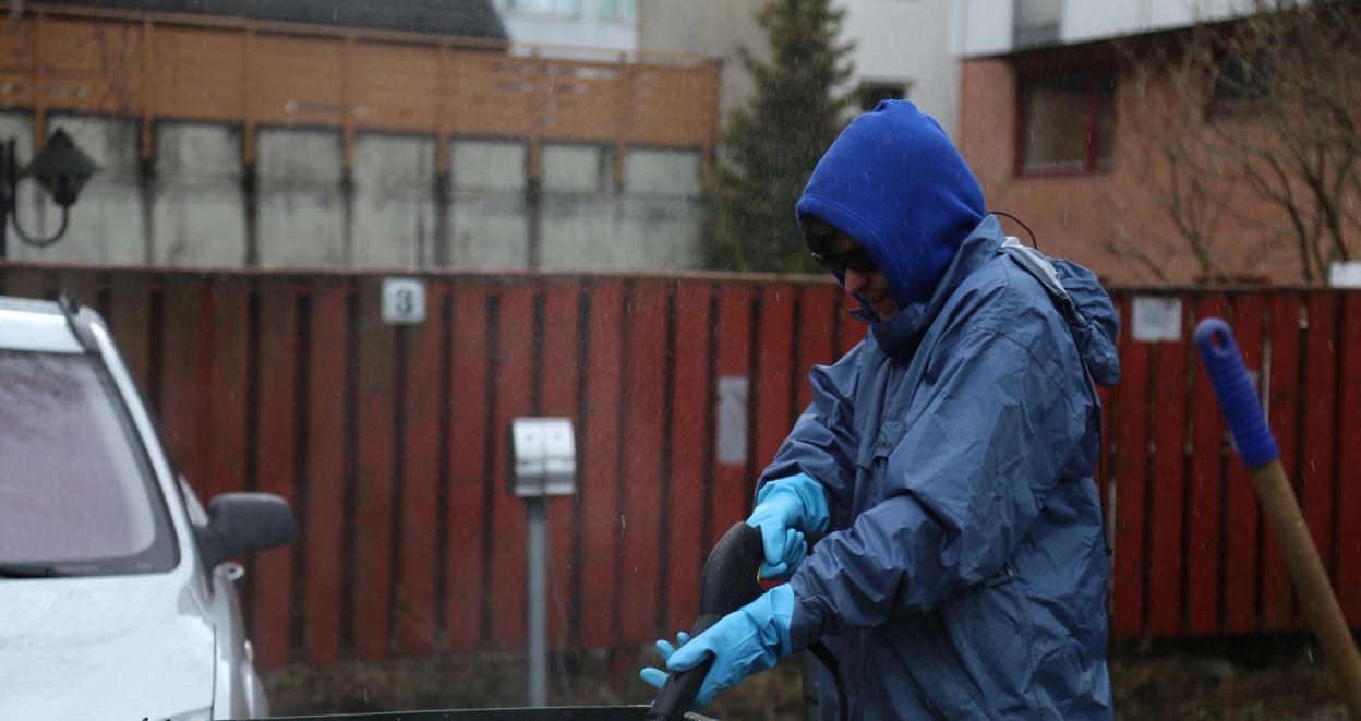 A person in a blue rain jacket and gloves power washes outdoor garbage bins in a residential area on a rainy day.