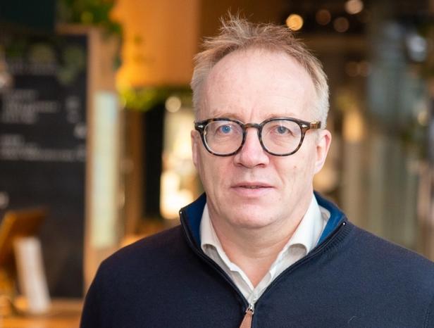 Jens Storli standing inside a warmly lit café or restaurant. He is wearing a navy blue sweater over a light-colored collared shirt and glasses. Behind him, a chalkboard menu, hanging wine glasses, and wooden furniture are visible, creating a cozy and inviting atmosphere.