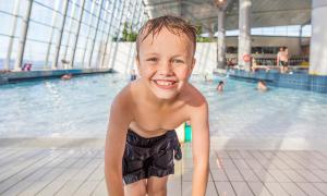 A smiling boy stands near the edge of a pool, wet from swimming, with a bright and cheerful expression. The indoor pool is visible in the background, with other people swimming and sunlight streaming through large windows.
