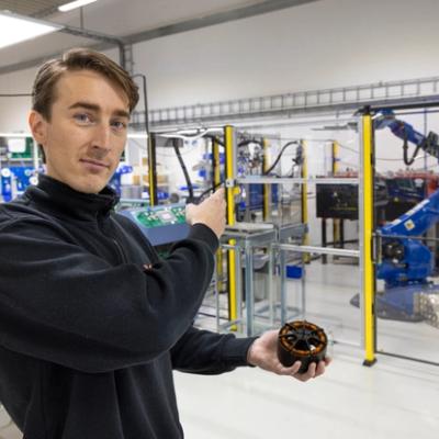 Alt text: A person stands in a factory with a robotic arm in the background, holding a small part and pointing at the machine.
