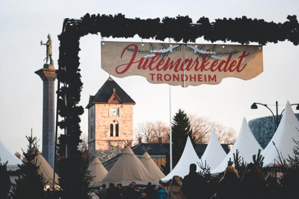 The entrance to "Julemarkedet Trondheim" (Trondheim Christmas Market) is framed by a festive arch decorated with greenery. A large banner with red lettering welcomes visitors. In the background, the historic Vår Frue Church and a statue atop a tall column are visible. White tents and traditional market stalls fill the square, with a crowd of warmly dressed people enjoying the holiday atmosphere. The winter sky and soft lighting add to the cozy, festive ambiance.