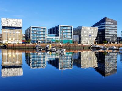 A panoramic view of modern, reflective buildings along a waterfront, with clear blue skies above. The glassy surface of the water perfectly mirrors the structures, enhancing the striking architecture of the buildings. The scene shows a mix of commercial and office spaces, with several boats docked along the harbor. The bright, sunny weather and the calm water create a clean, contemporary urban atmosphere.