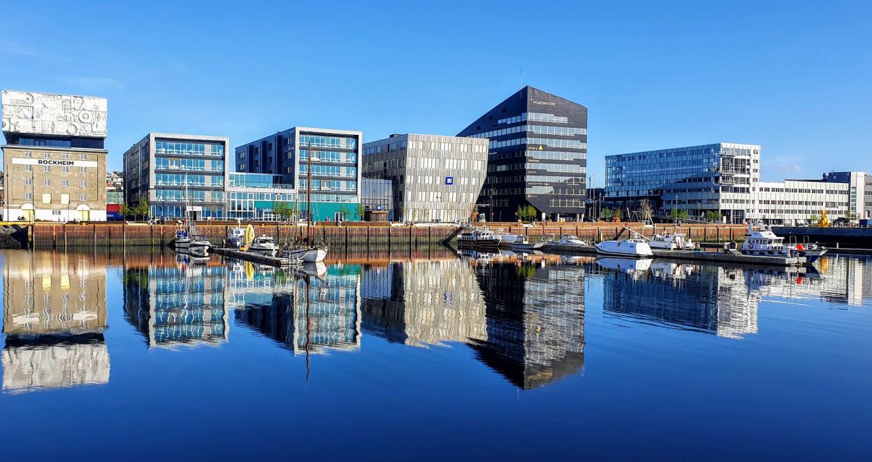 A panoramic view of modern, reflective buildings along a waterfront, with clear blue skies above. The glassy surface of the water perfectly mirrors the structures, enhancing the striking architecture of the buildings. The scene shows a mix of commercial and office spaces, with several boats docked along the harbor. The bright, sunny weather and the calm water create a clean, contemporary urban atmosphere.