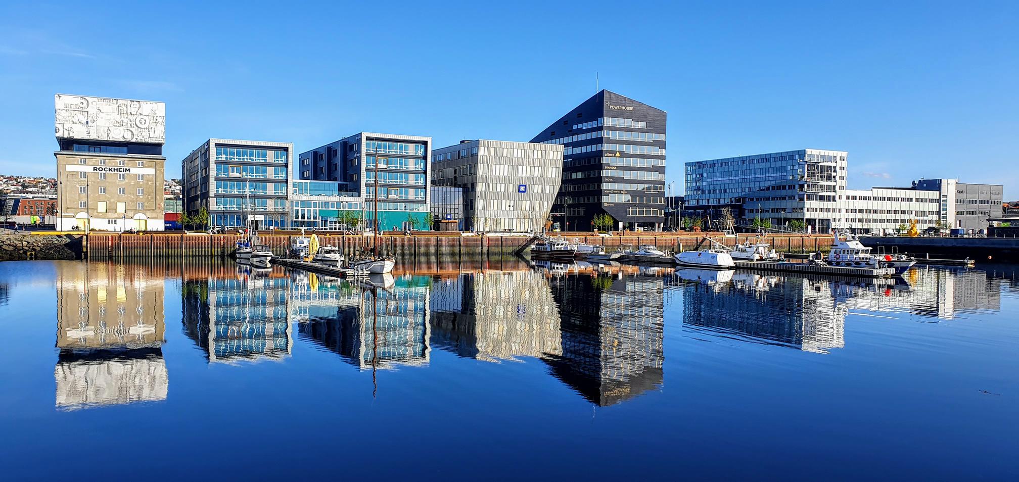 A panoramic view of modern, reflective buildings along a waterfront, with clear blue skies above. The glassy surface of the water perfectly mirrors the structures, enhancing the striking architecture of the buildings. The scene shows a mix of commercial and office spaces, with several boats docked along the harbor. The bright, sunny weather and the calm water create a clean, contemporary urban atmosphere.