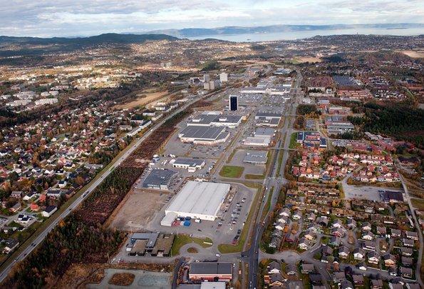 An aerial view of the Tiller area in Trondheim, featuring a mix of industrial buildings, shopping centers, and residential neighborhoods. In the foreground, the layout of streets and buildings is clearly visible, with green areas in the background