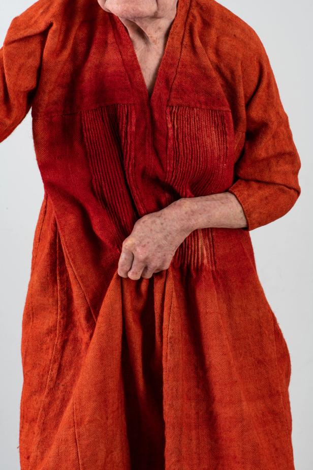 Close-up of a medieval woman wearing an orange-red robe with intricate pleats, holding the fabric with one hand.