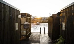 A wooden walkway between two dark wooden structures leads to a small pier by the water, with the sun setting in the background. A small boat floats on the calm water, and buildings with industrial elements line the opposite shore. Signs with symbols and text are mounted on the wooden walls, suggesting this might be a sauna or bathing facility. The warm glow of the sunset casts long shadows, creating a serene atmosphere.