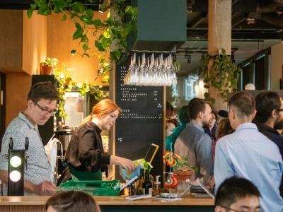 A lively café or bar in Trondheim filled with people socializing. A bartender serves drinks while guests enjoy the vibrant atmosphere, surrounded by greenery and modern decor.
