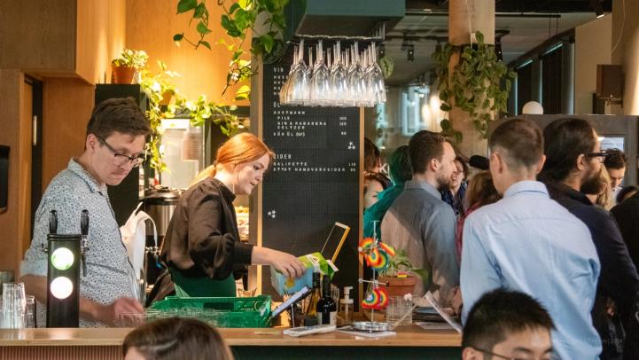 A lively café or bar in Trondheim filled with people socializing. A bartender serves drinks while guests enjoy the vibrant atmosphere, surrounded by greenery and modern decor.