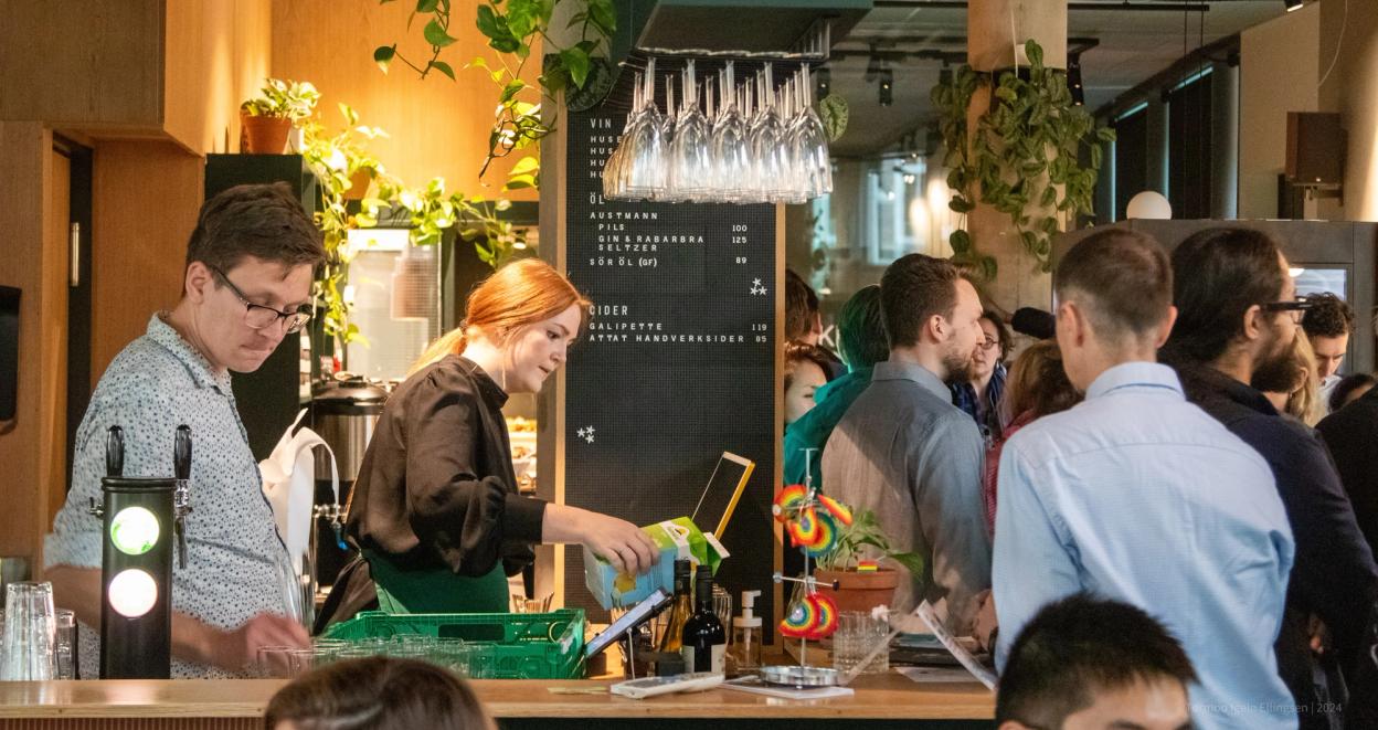 A lively café or bar in Trondheim filled with people socializing. A bartender serves drinks while guests enjoy the vibrant atmosphere, surrounded by greenery and modern decor.