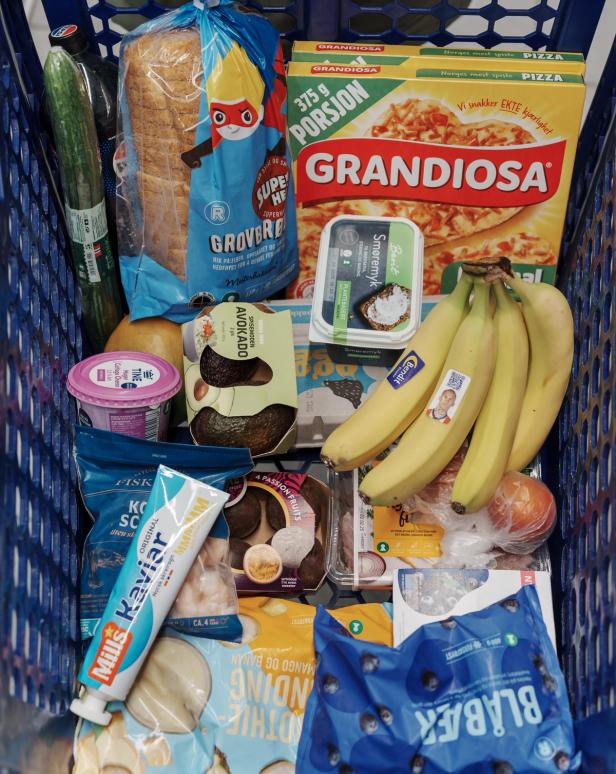 A shopping cart filled with groceries including bread, pizza (Grandiosa), bananas, yogurt, cucumbers, a tube of mustard, cookies, and other items.