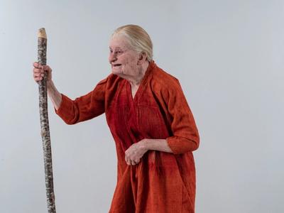 This is a remake of a medieval woman, depicted with an orange tunic and holding a walking stick, showcasing an aged appearance.