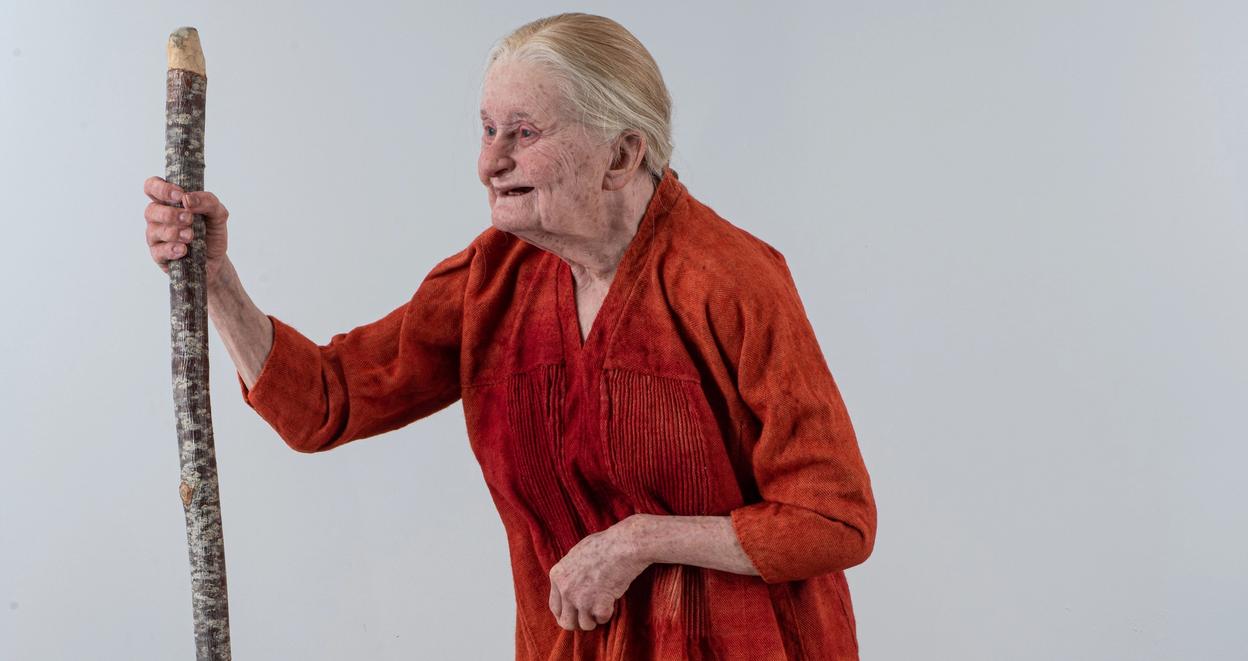 This is a remake of a medieval woman, depicted with an orange tunic and holding a walking stick, showcasing an aged appearance.