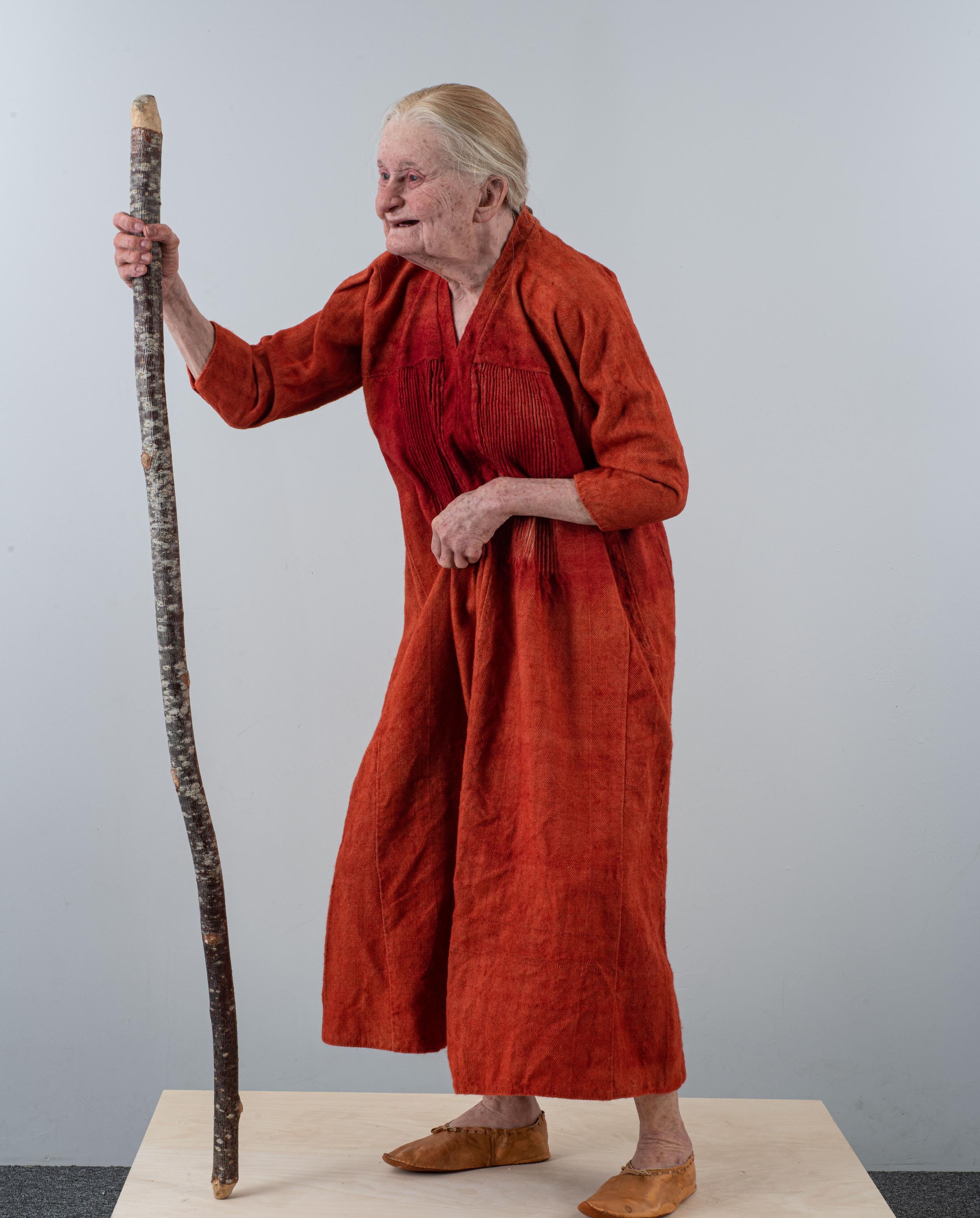 This is a remake of a medieval woman, depicted with an orange tunic and holding a walking stick, showcasing an aged appearance.