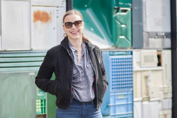 This image shows a person standing with a smile, wearing sunglasses and a black jacket, in front of a wall covered with various colored panels or doors. The background is a vibrant mix of shades of green, blue, and other colors. The person appears confident and cheerful.