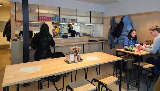 Inside of a restaurant selling ramen. The bossman is taking orders and a young couple is enjoying a nice lunch