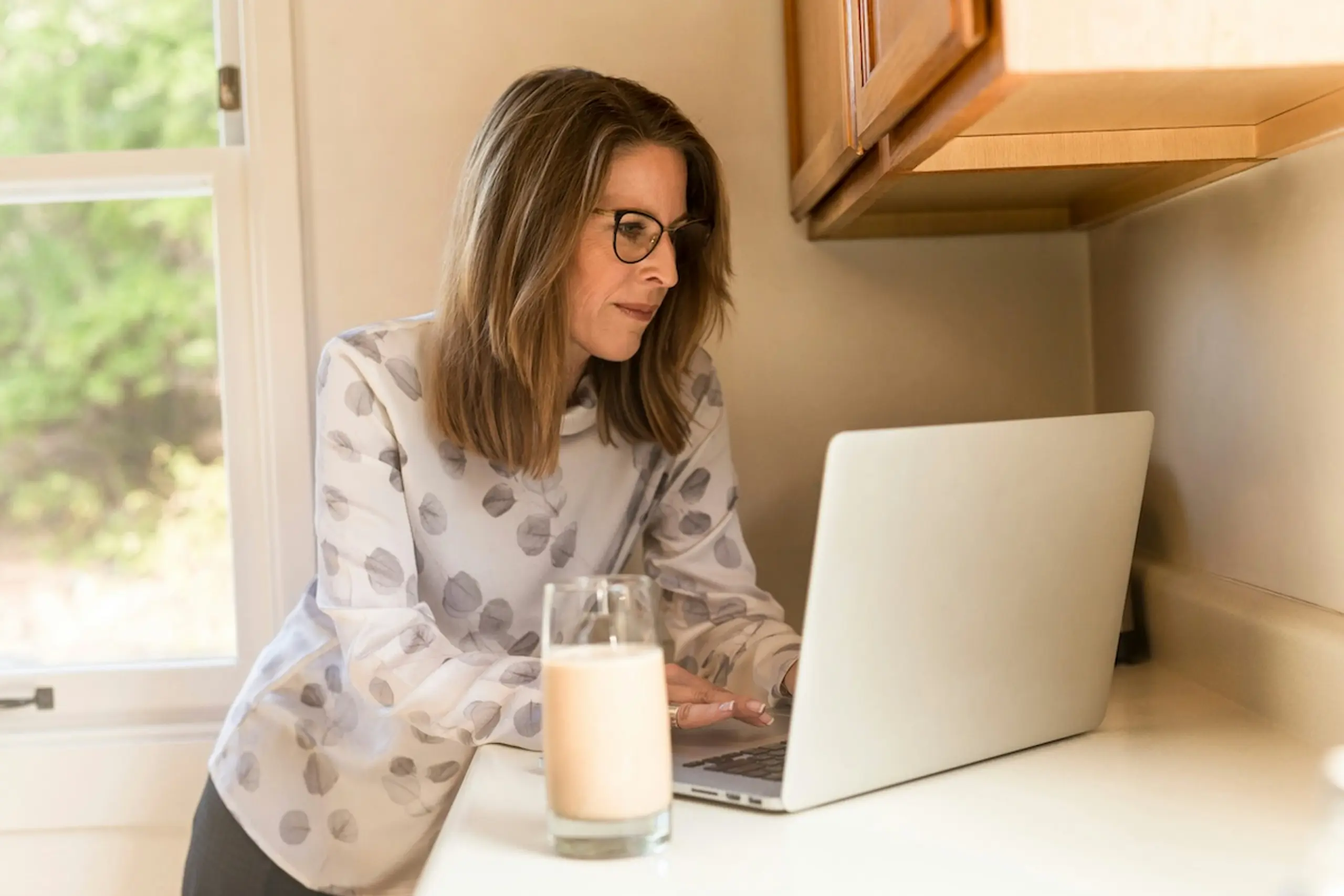 woman wearing glasses looking at laptop on counter next to a glass of milk