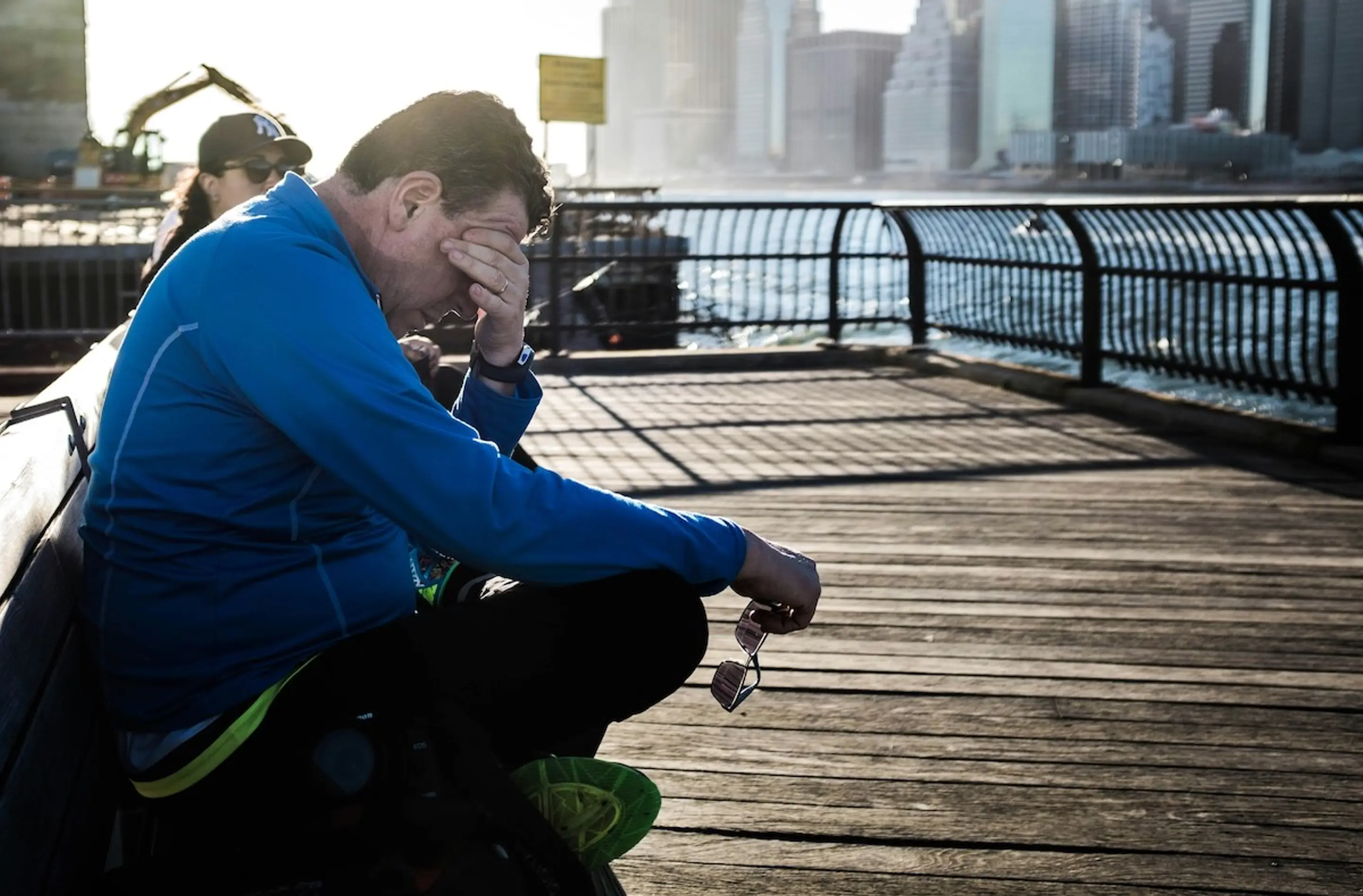A man covering his eyes while sitting on a bench by the river