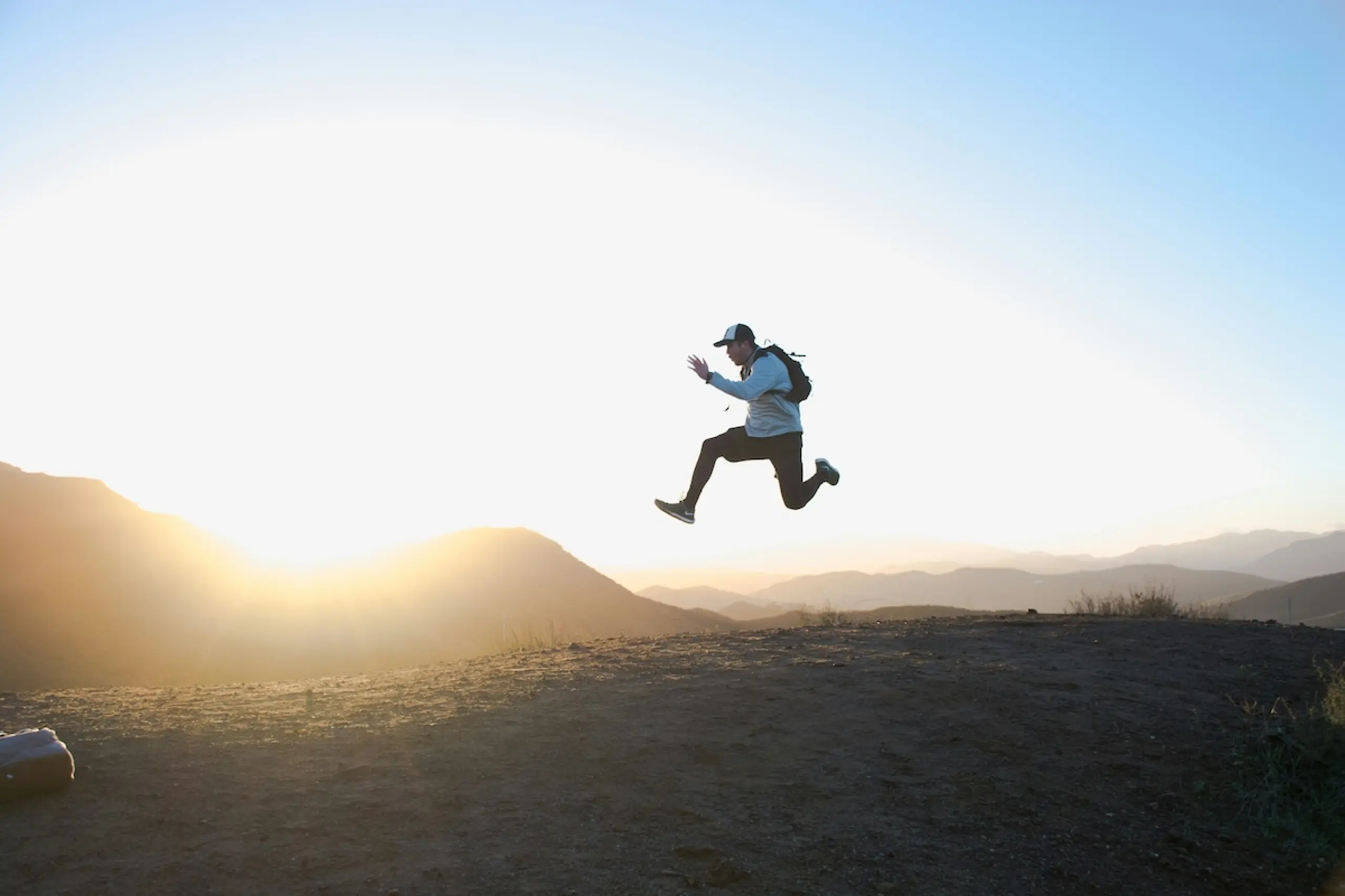 man wearing backpack and hat on mountain top jumping in the air with the sun in the background