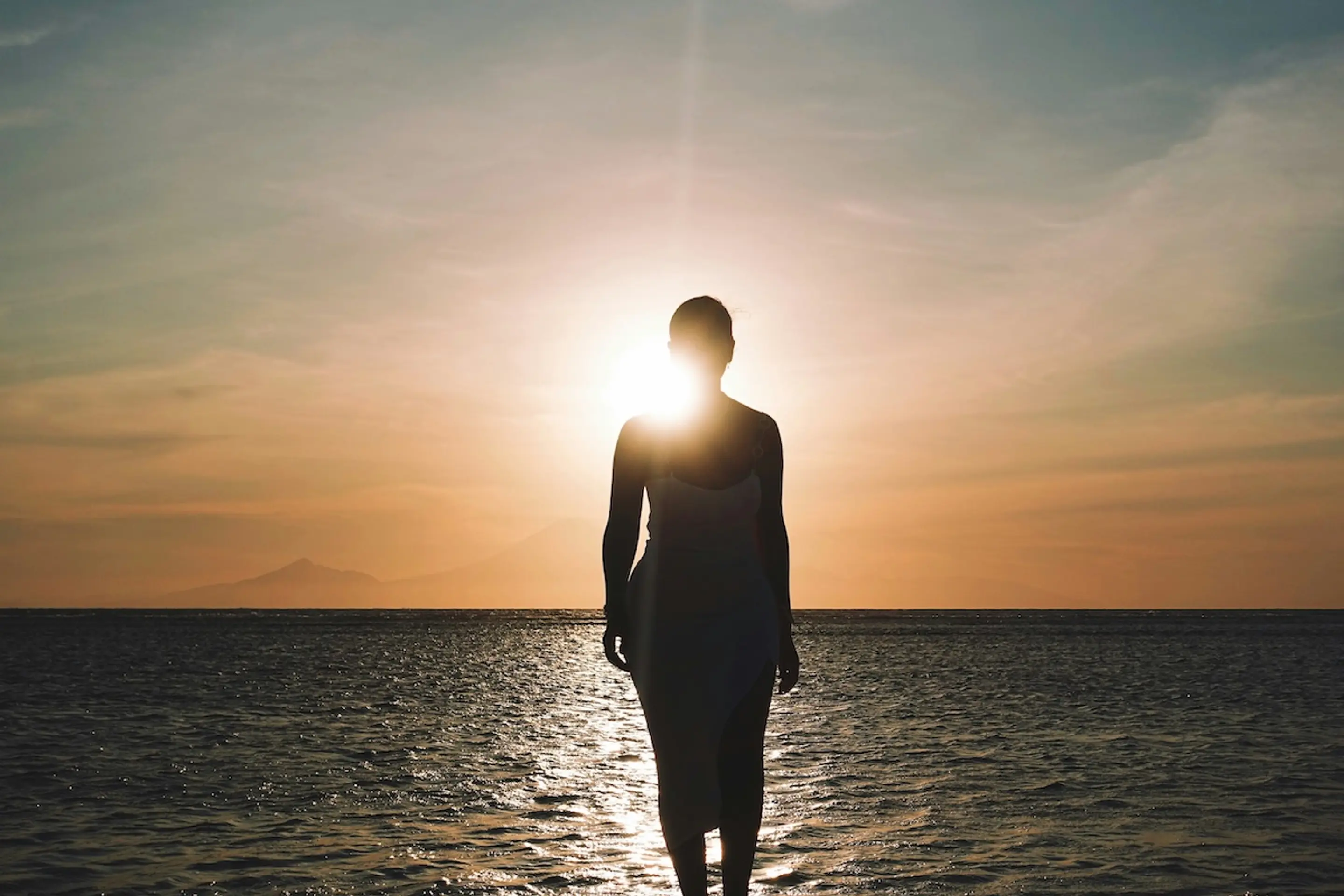 person standing by water looking at sun