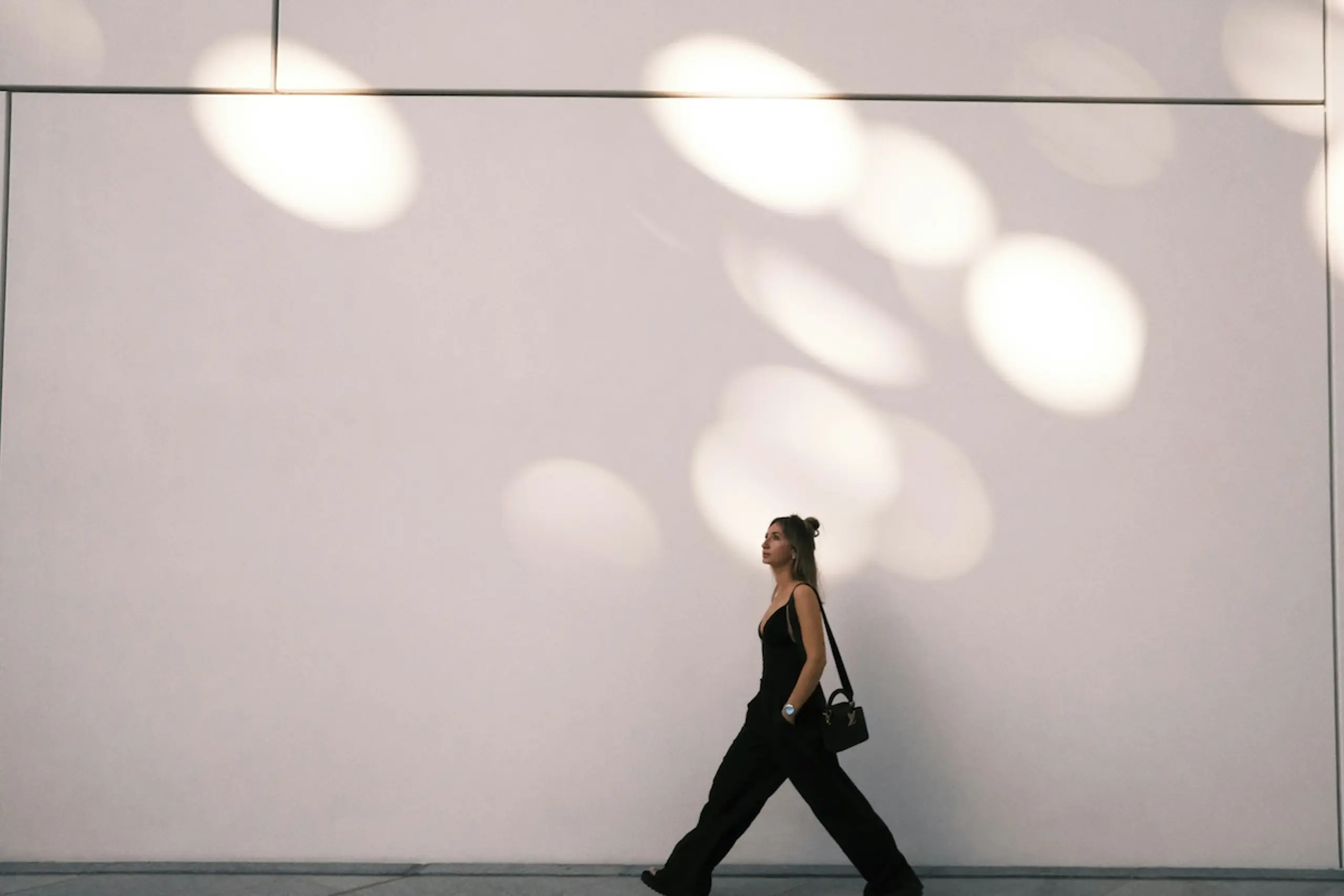 woman walking in front of white wall with purse and hands in pockets