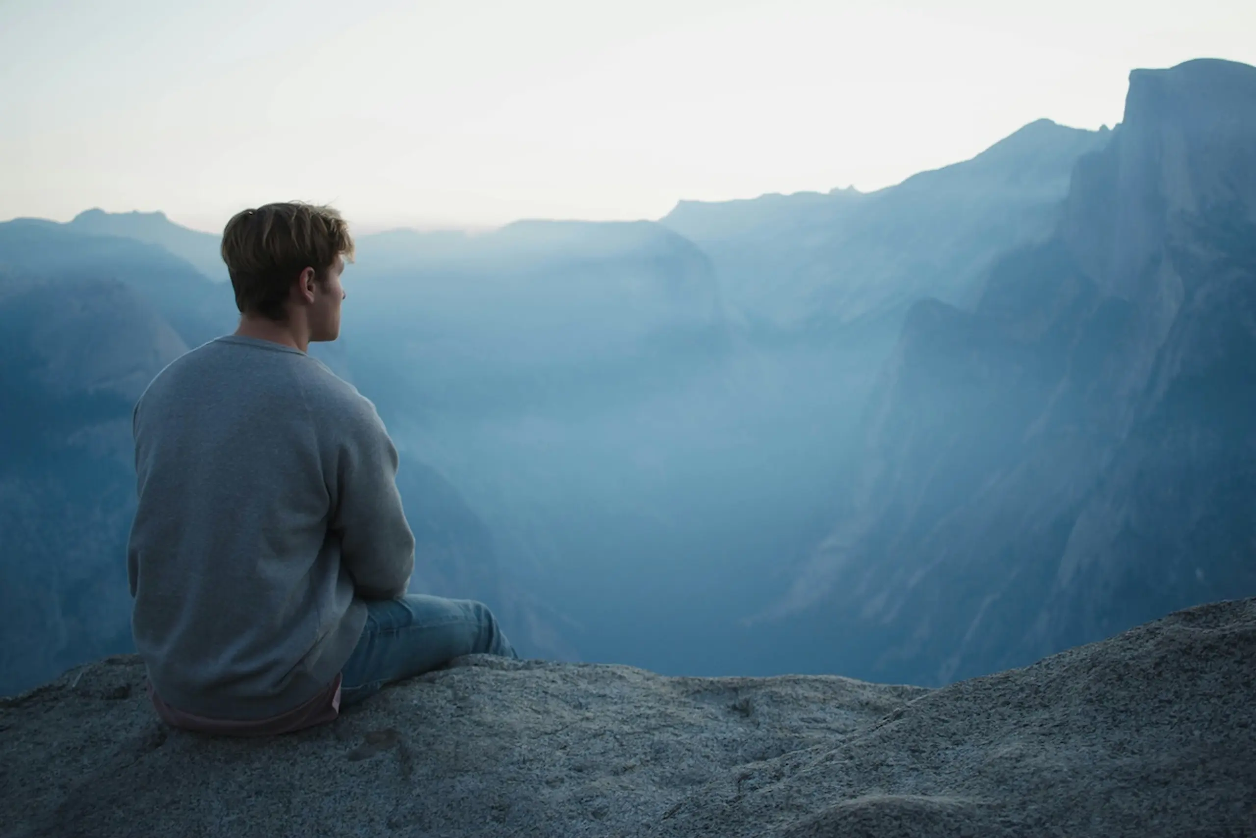 man in sweatshirt and pants sitting on mountainside looking at mountain range