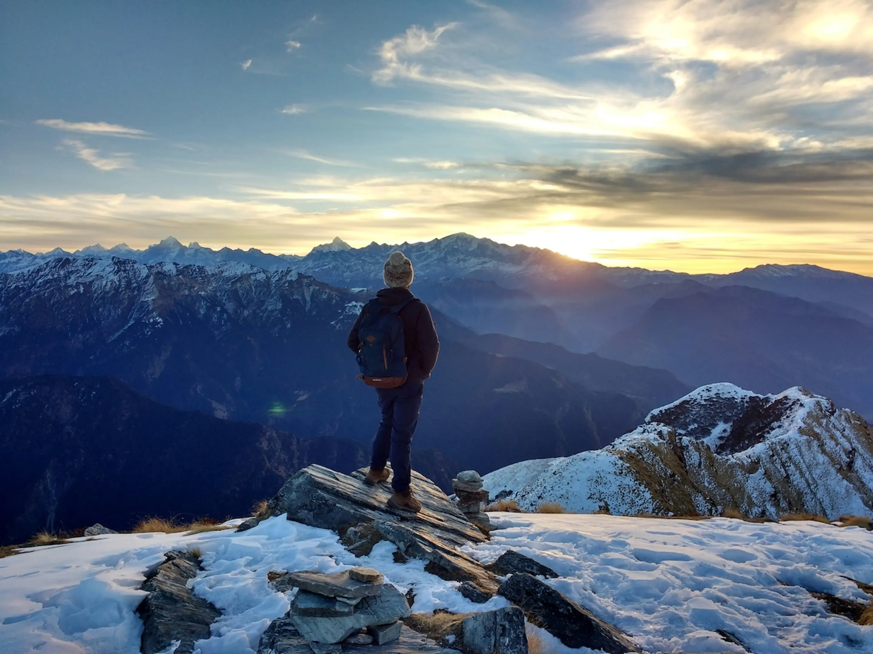 person standing on mountain peak overlooking snowy mountains during sunset