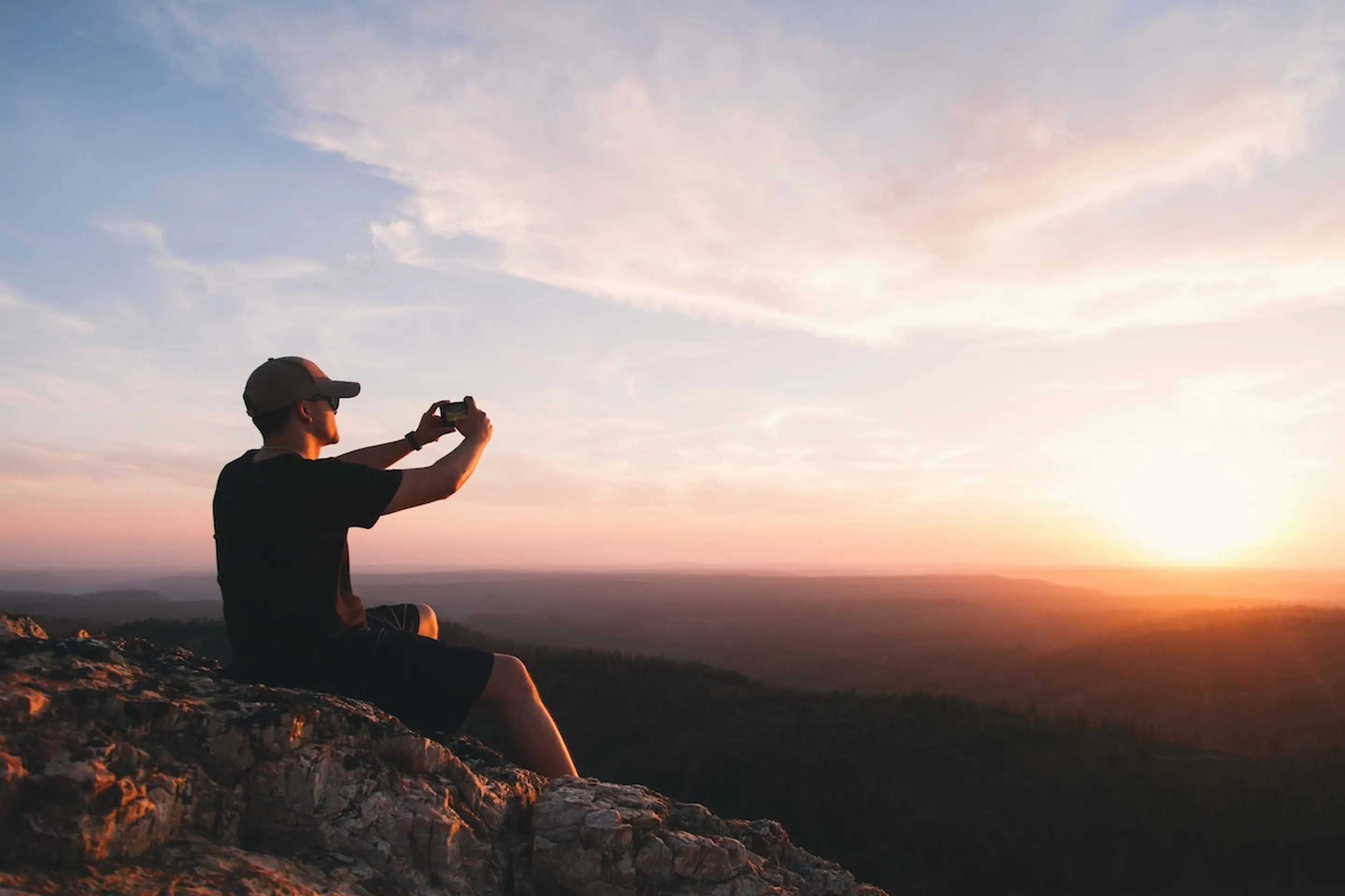 man sitting on rock taking picture of sunset