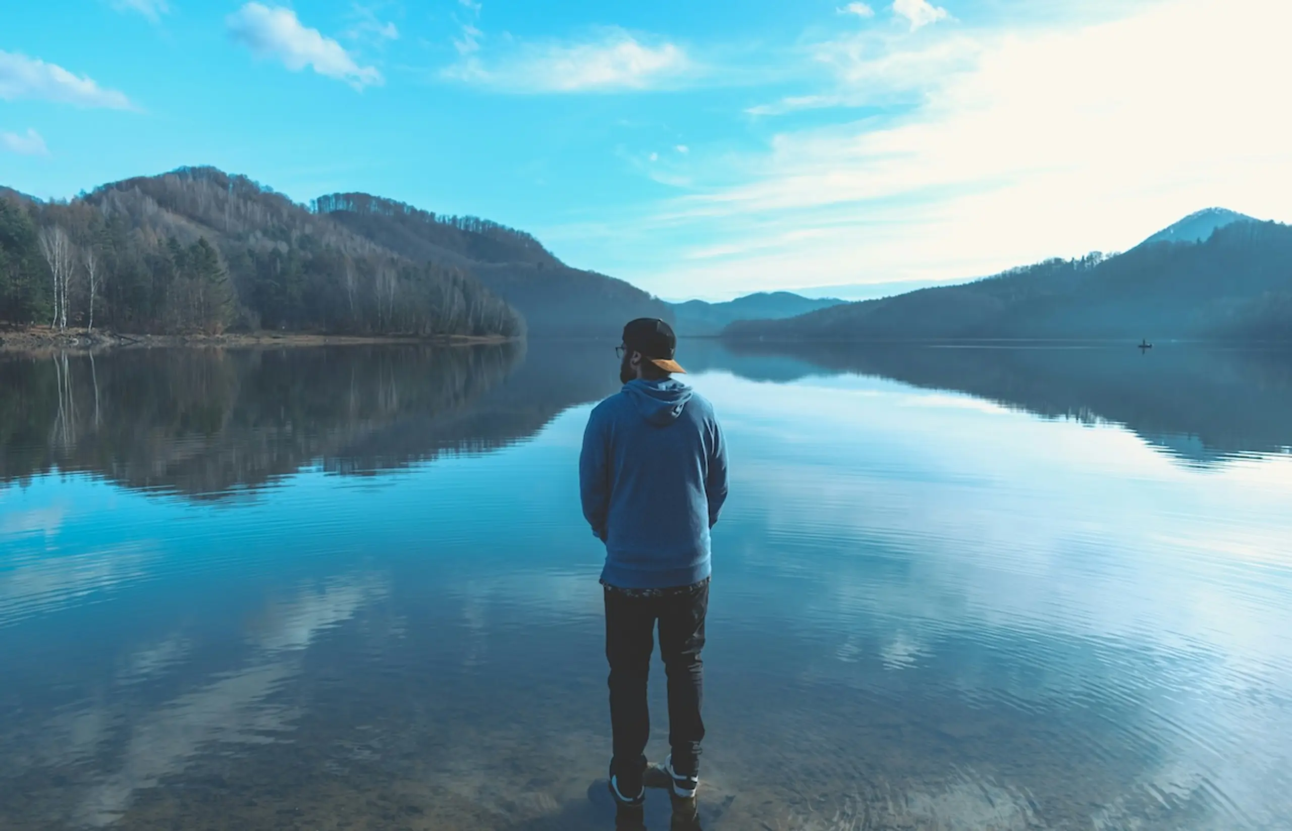 man standing in front of water and mountains with hat on backwards