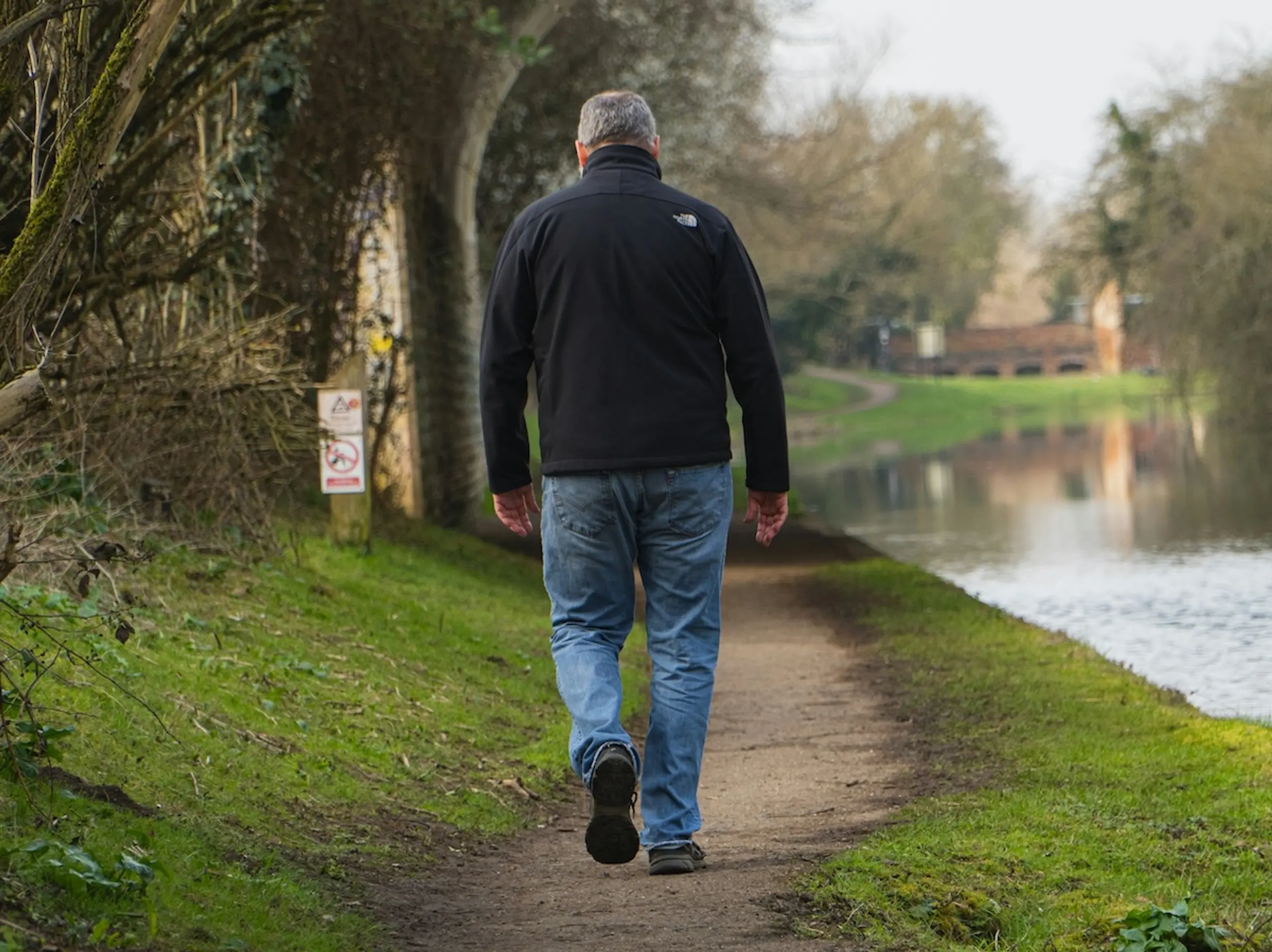 man walking on path among trees with a body of water to his right