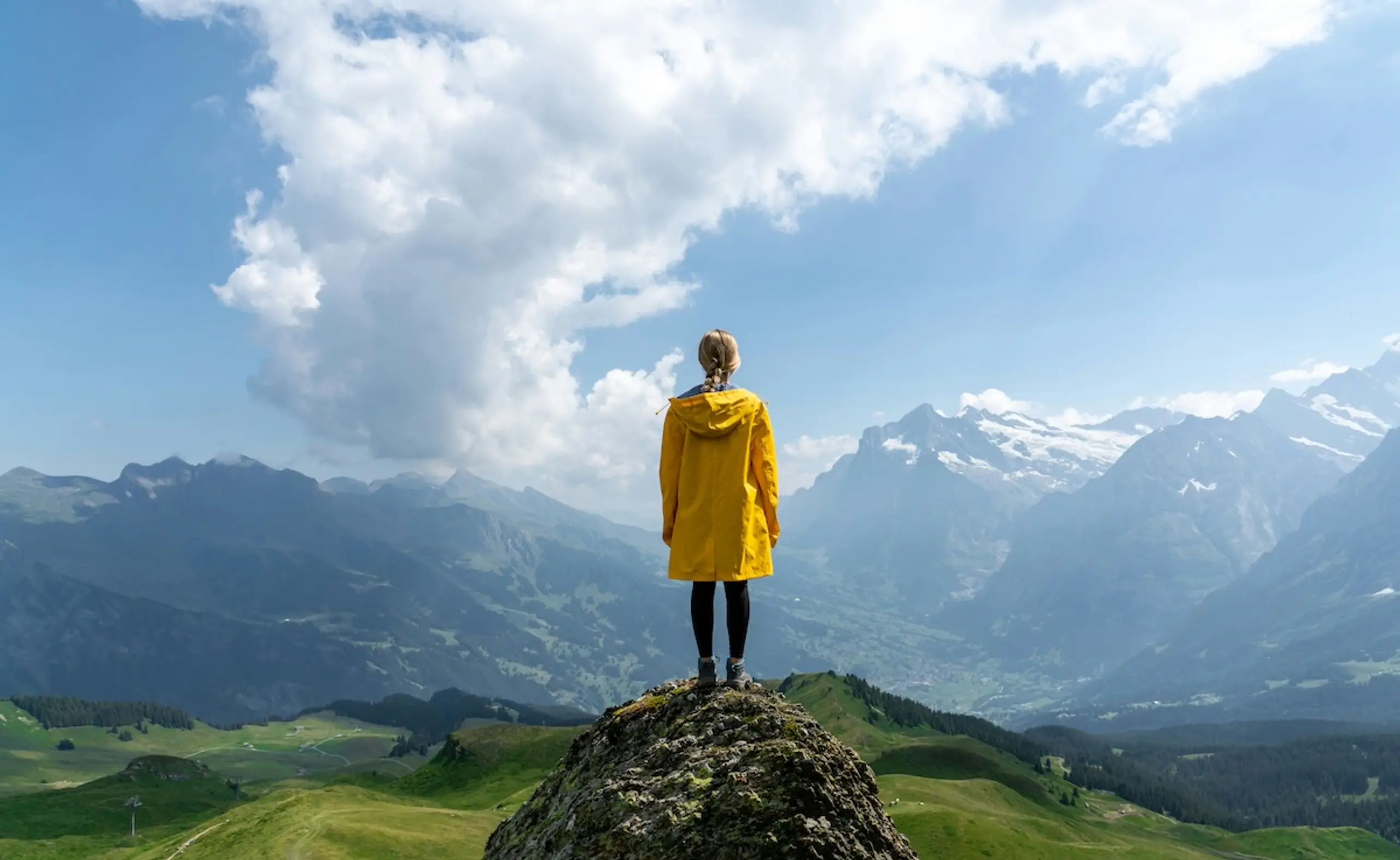 woman in yellow jacket standing on mountain looking at snow-covered mountain range and clouds