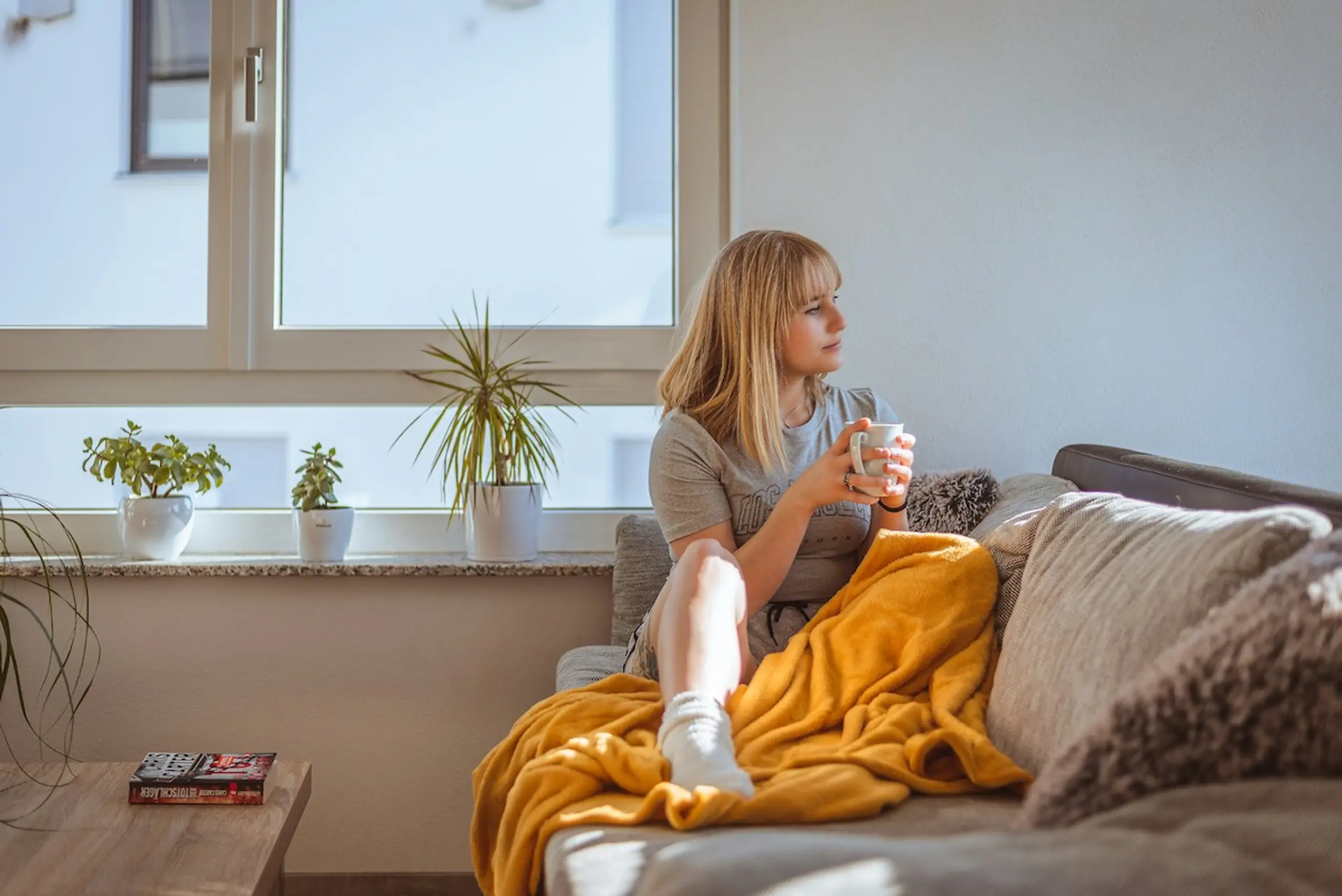 A woman with a cup of coffee on her couch at home looking out the window