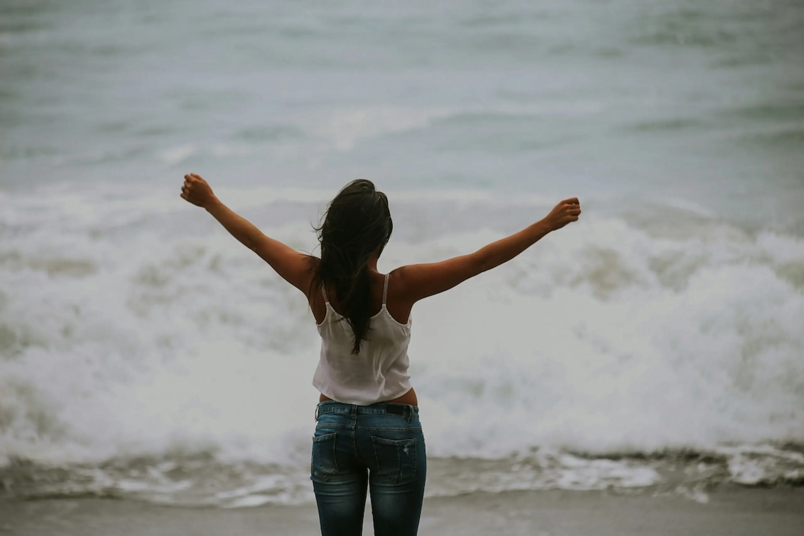 woman wearing jeans and white tank top with open arms standing in front of crashing wave at shoreline
