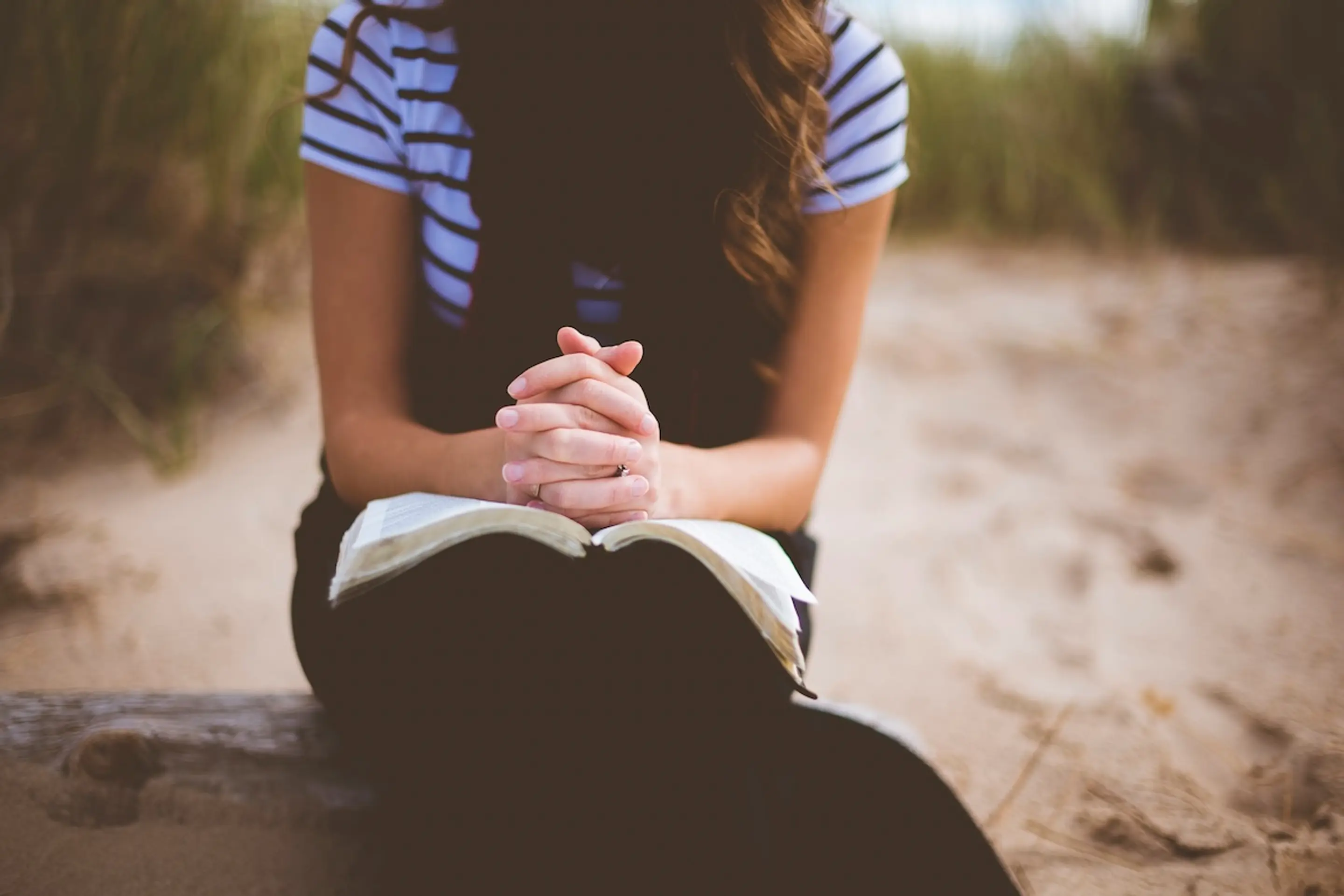 woman sitting on beach with book in lap and hands clasped