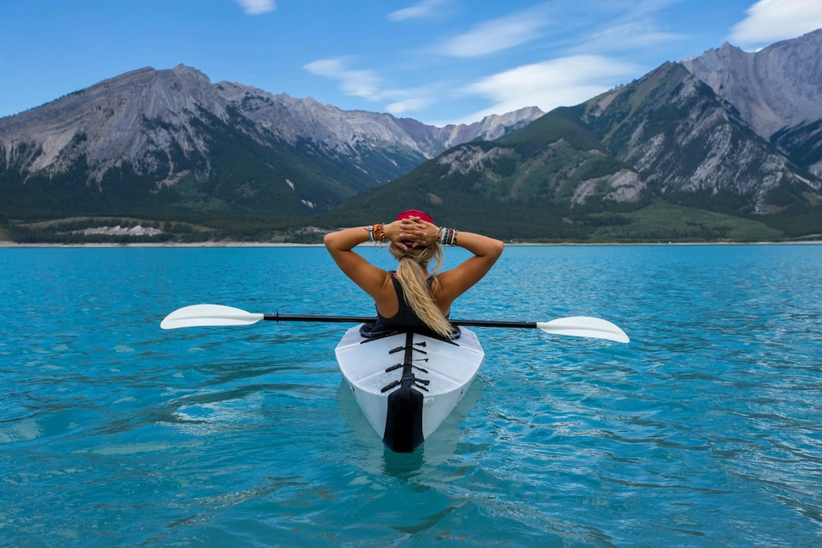 woman with blonde ponytail in kayak in blue water looking at mountains with hands on her head
