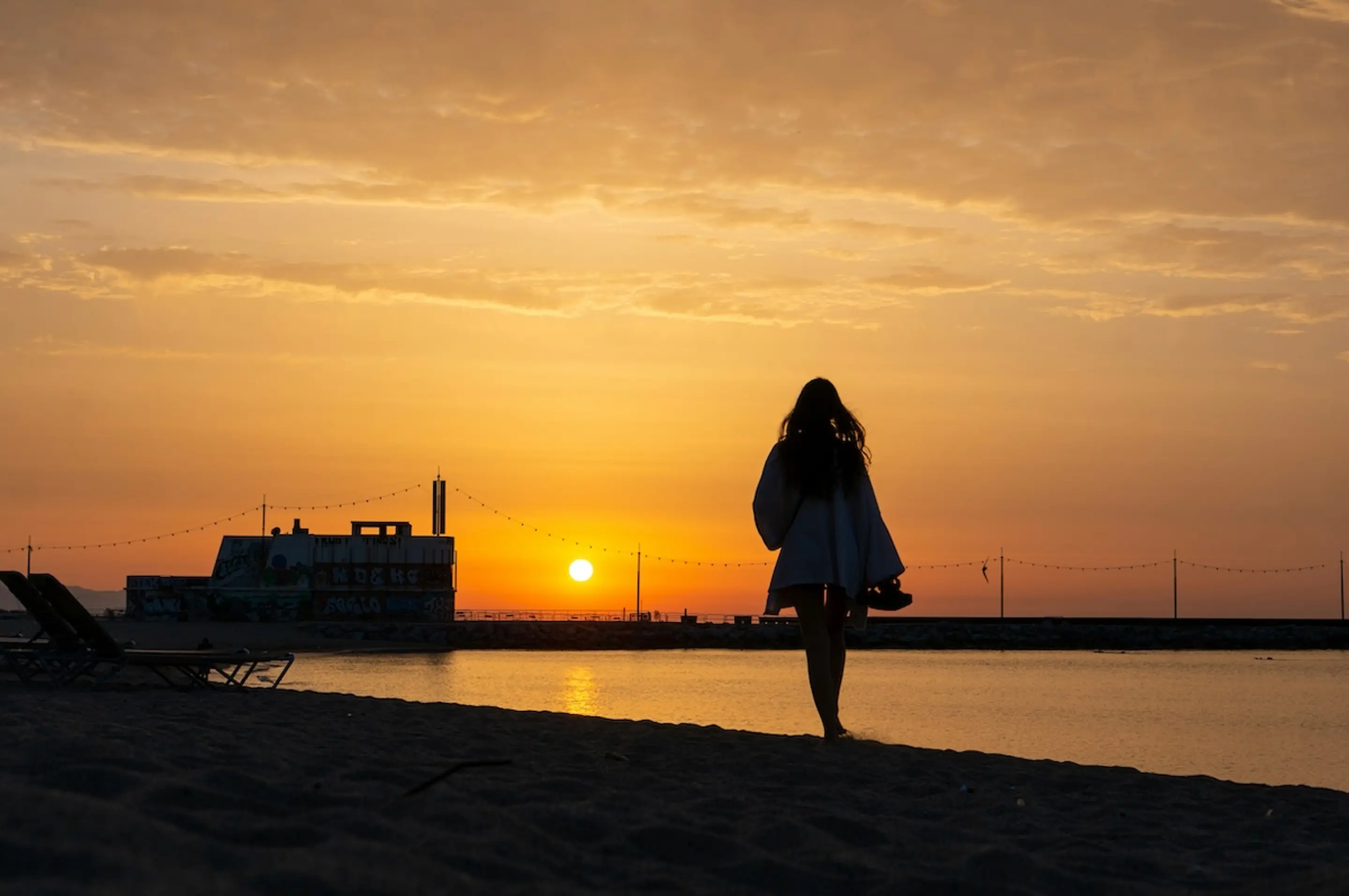 woman walking on beach at sunset carrying shoes by the water