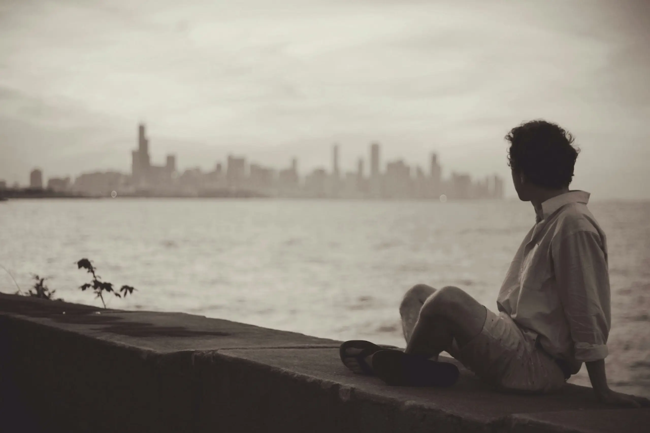 person sitting cross-legged on wall next to water looking at city skyline across water