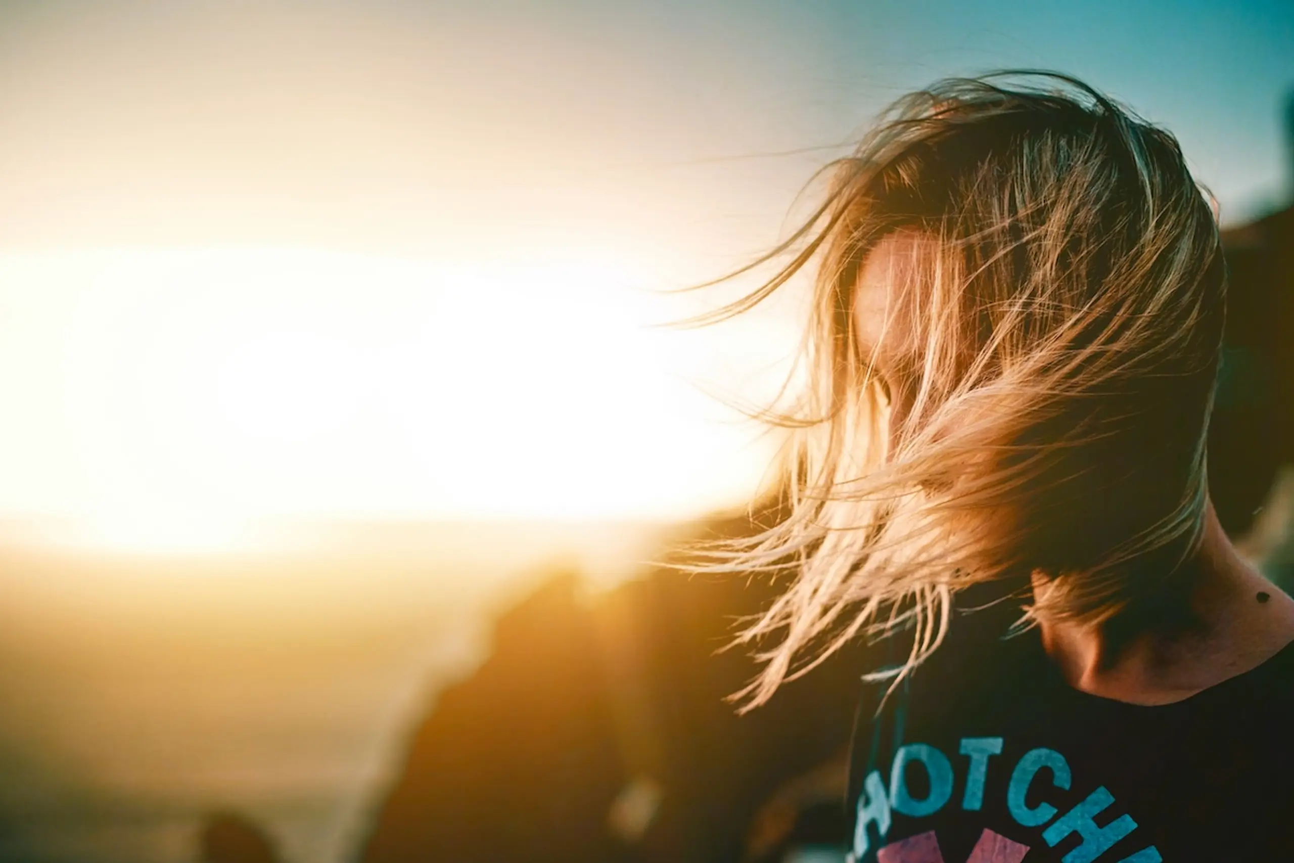 person with blonde hair blowing in the wind on mountainside with sunset in the background