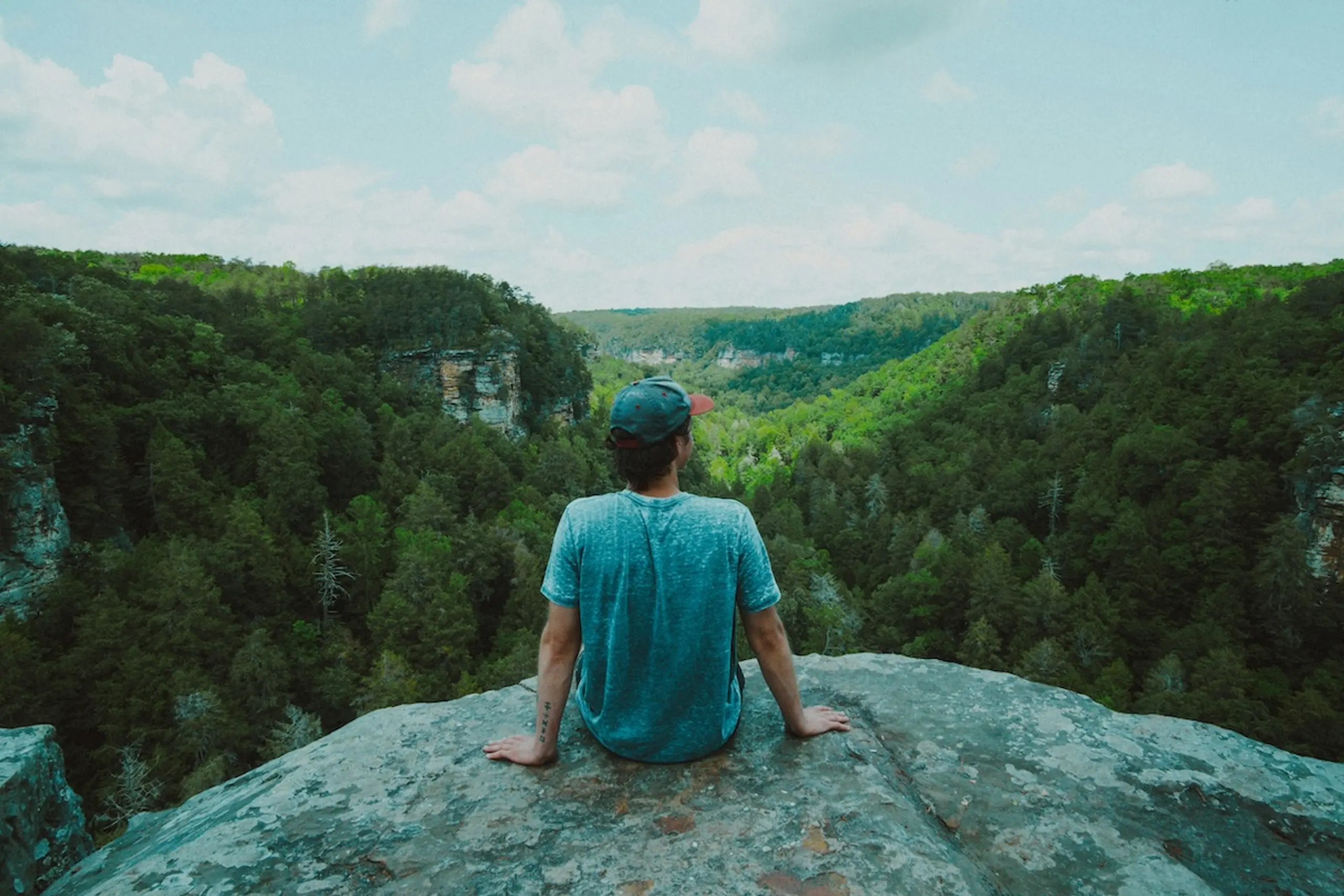 person sitting on rock wearing hat and t-shirt overlooking trees and hills in the distance