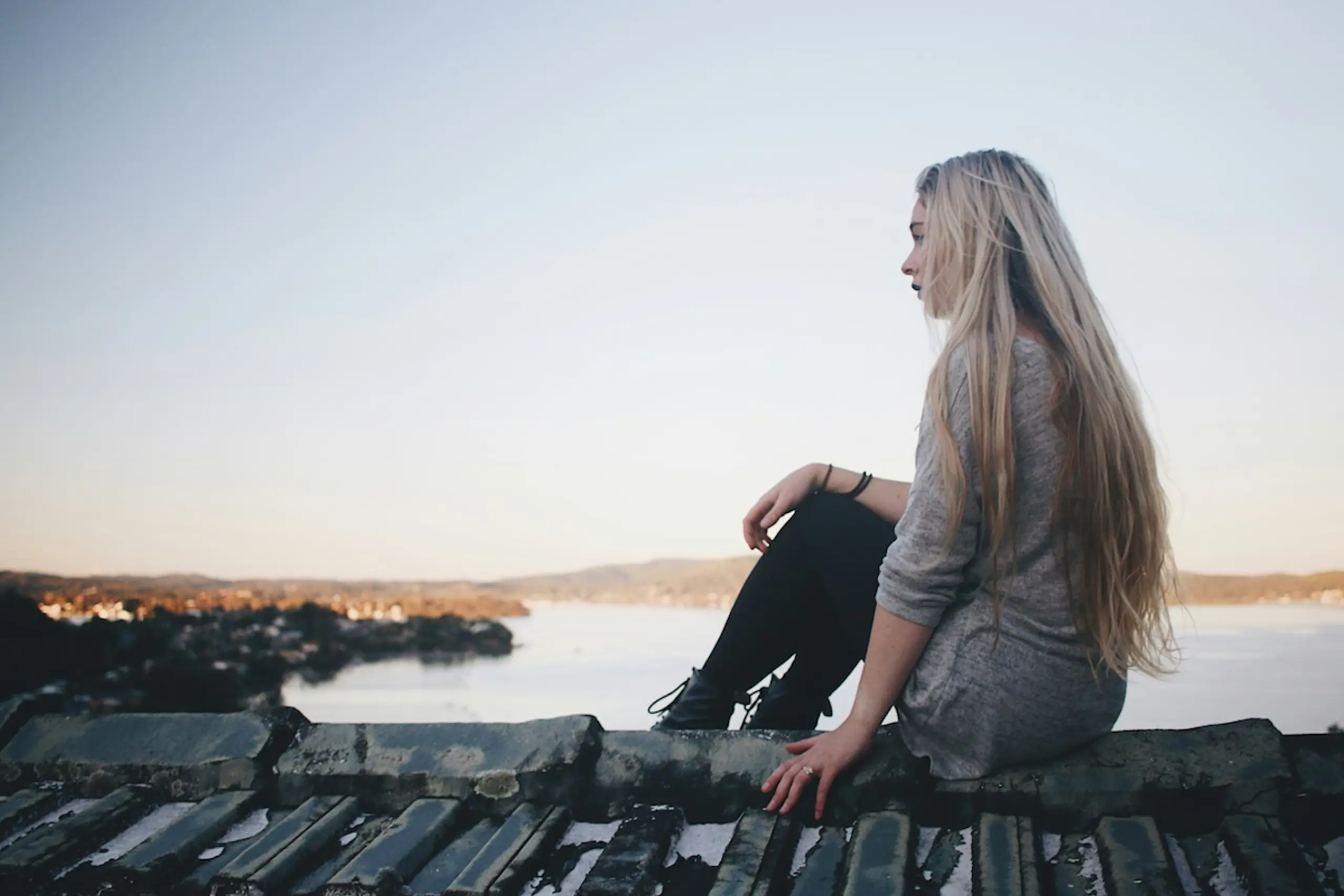 woman sitting on dock looking at town on water's edge with hills in the background