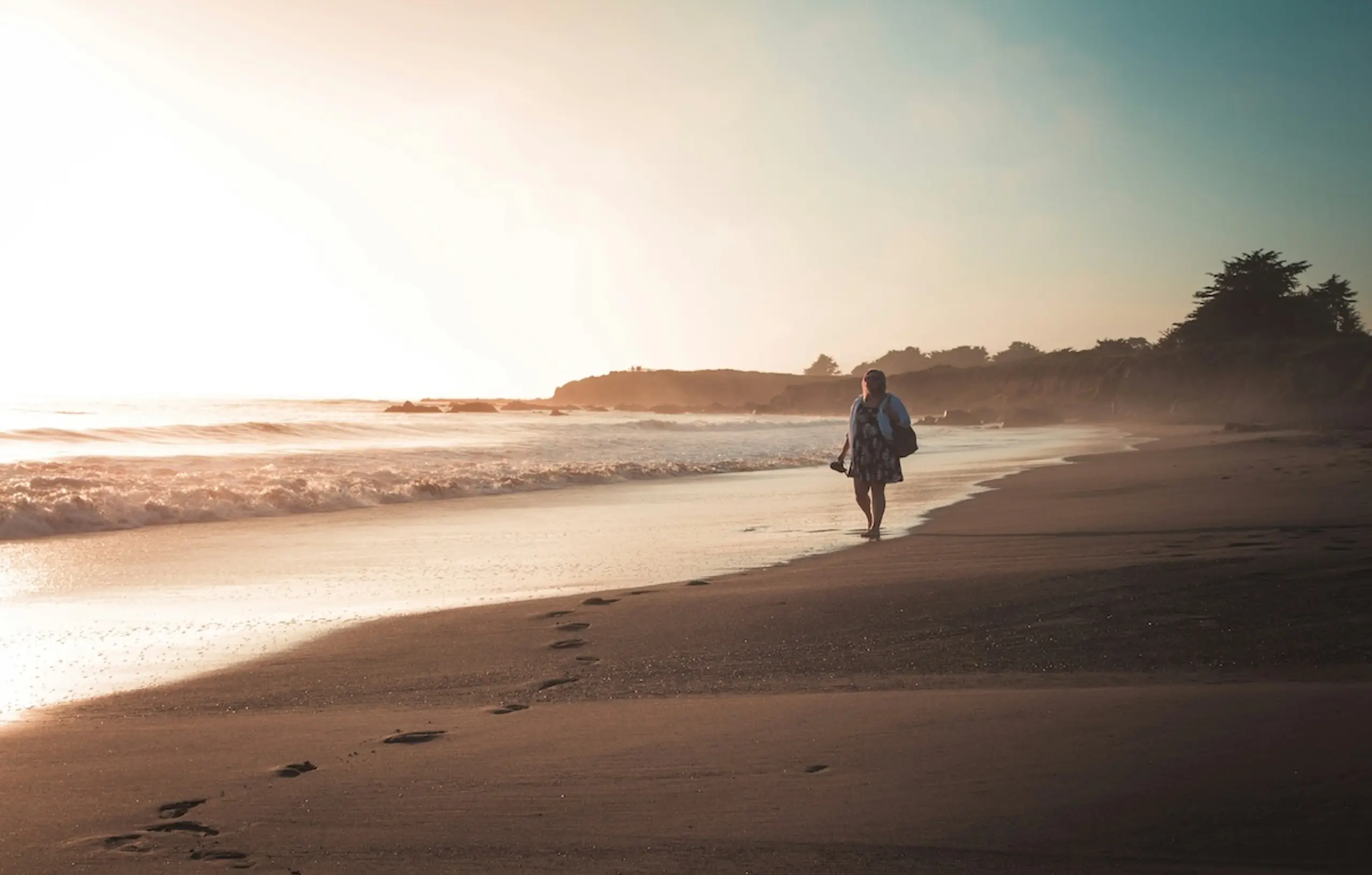 person walking down beach looking at water carrying sandals
