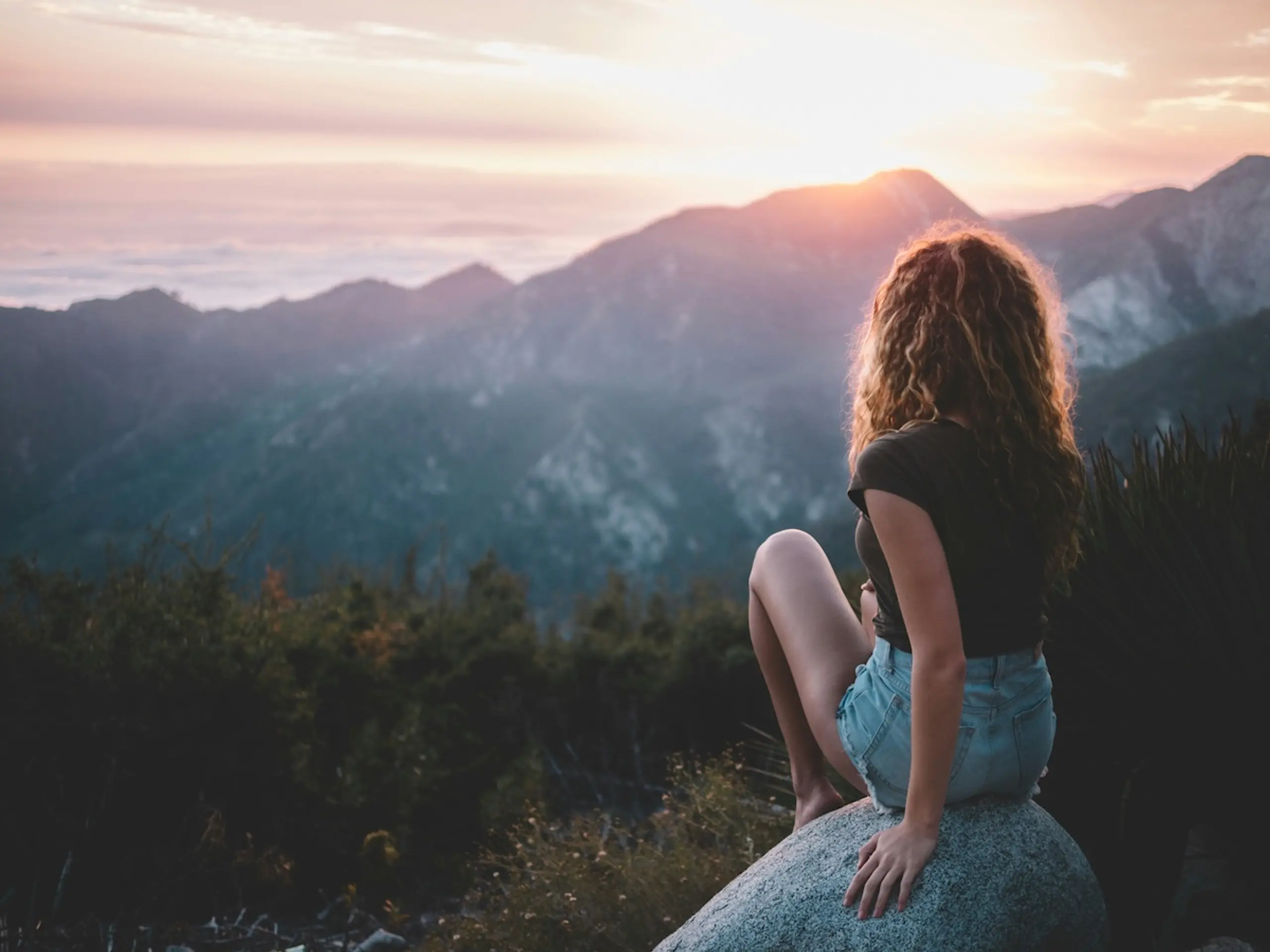 woman in shorts and t-shirt sitting on a rock watching sunset over mountains
